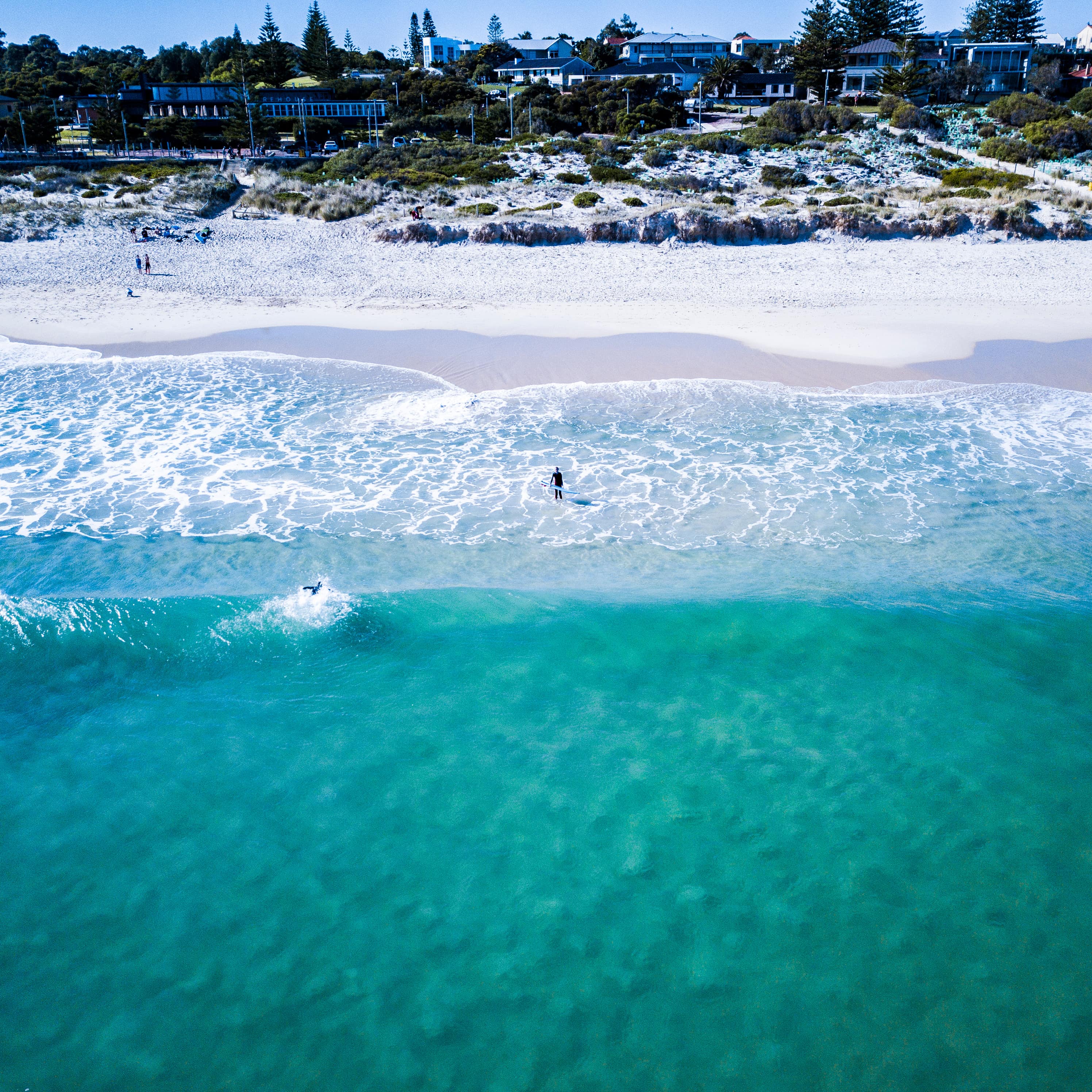Aerial view of a man in the shallow water at the beach holding a surfboard and another surfer catching a wave