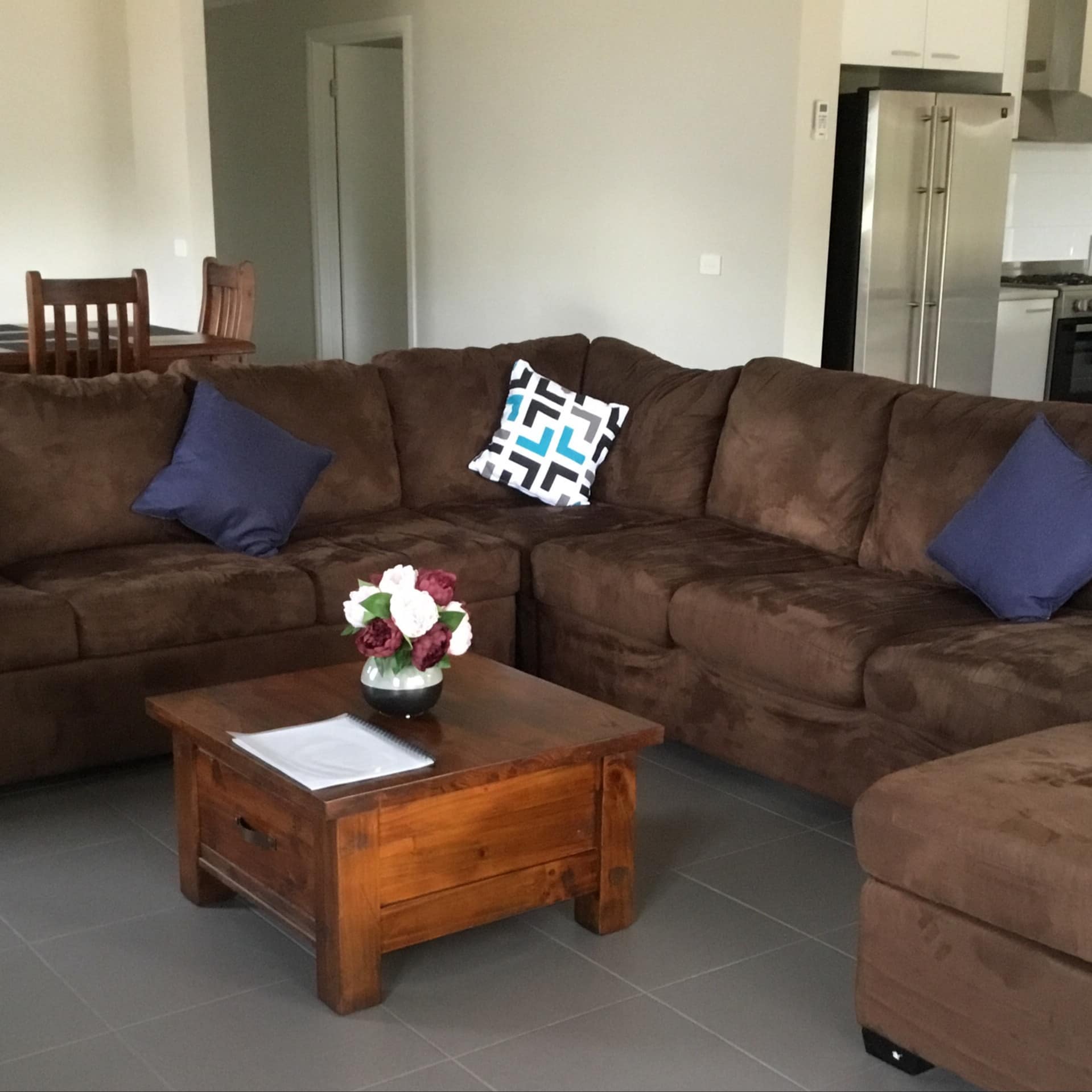 An open living room and kitchen with grey tile floors, a brown suede sectional, wooden coffee table, and wood dining set.